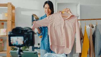 ung asiatisk dammodedesigner som använder mobiltelefon tar emot inköpsorder och visar kläder som spelar in video live streaming online med kamera. småföretagare, online marknadsföringskoncept.