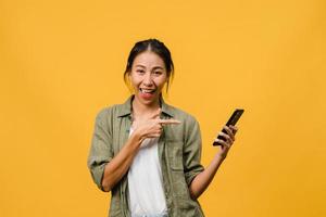 ung asiatisk dam som använder mobiltelefon med glada uttryck, visar något fantastiskt på tomt utrymme i vardagsduk och tittar på kameran isolerad över gul bakgrund. ansiktsuttryck koncept. foto