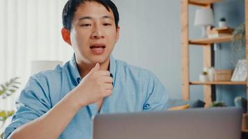 ung asiatisk affärsman som använder bärbar dator pratar med kollegor om planering i videosamtal medan smart arbetar hemifrån i vardagsrummet. självisolering, social distansering, karantän för förebyggande av corona-virus.