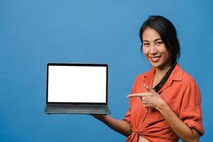 ung asiatisk dam visar tom bärbar datorskärm med positivt uttryck, ler brett, klädd i vardagskläder som känner lycka isolerad på blå bakgrund. dator med vit skärm i kvinnlig hand. foto