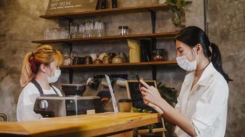 unga asien kvinnliga bär ansiktsmask självbetjäning använda mobiltelefon betala kontaktlös på restaurang. flicka barista talar ta emot beställning från kund på café. livsstil nytt normalt efter corona -virus koncept. foto