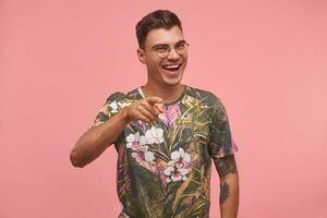 ung härlig man i en blommig t-shirt skrattande och pekande till de kamera, höjning index finger, bär glasögon och tryckt t-shirt, stående över rosa bakgrund foto