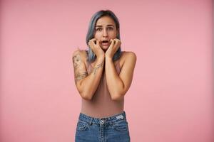 skrämd ung attraktiv tatuerade kvinna med kort blå hår innehav henne ansikte med Uppfostrad händer och ser rädd på kamera, isolerat över rosa bakgrund foto