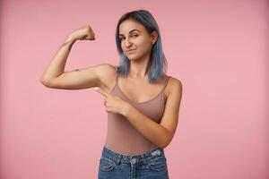 porträtt av ung attraktiv tatuerade kvinna med kort frisyr pekande på henne Uppfostrad hand medan som visar henne stark biceps, stående över rosa bakgrund foto