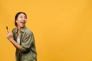 ung asiatisk dam med telefon med positivt uttryck, ler brett, klädd i vardagskläder som känner lycka och står isolerad på gul bakgrund. glad förtjusande glad kvinna jublar över framgång.