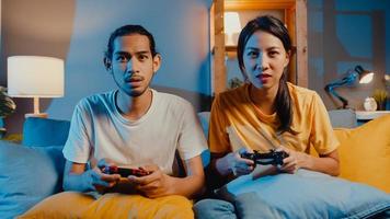 lyckligt asien ungt par man och kvinna sitter i soffan använda joystick -kontroller spela videospel tillbringa rolig tid tillsammans i vardagsrummet på natten. asiatiska gift par familj livsstil, stanna hemma koncept. foto