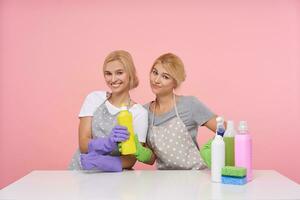 inomhus- Foto av ung vithuvud systrar med tillfällig frisyr framställning ha sönder med rengöring och Sammanträde över rosa bakgrund med flerfärgad flaskor av tvättmedel