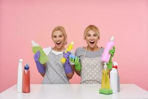 glad ung vithuvud rengöring damer innehav flaskor med hushåll kemikalier och ser lyckligt på kamera med öppnad munnar, isolerat över rosa bakgrund foto