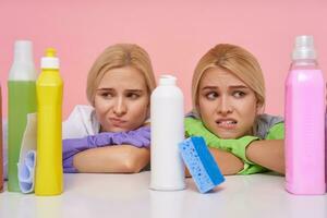 hemskt ung brunögd blond rengöring damer innehav deras händer på bänkskivan och ser tröttsamt på flaskor med hushåll kemikalier, isolerat över rosa bakgrund foto