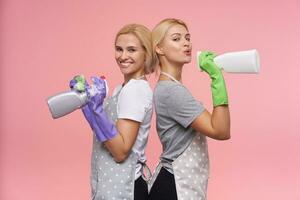 positiv ung attraktiv vithuvud rengöring damer bär sudd handskar medan rengöring hus och innehav spray flaskor, stående över rosa bakgrund foto