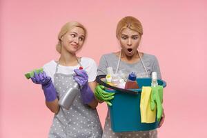 förvirrad ung vithuvud rengöring damer bär arbetssätt kläder och sudd handskar medan stående över rosa bakgrund med flaskor av tvättmedel och hushåll hink foto