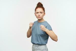 porträtt av allvarlig rödhårig flicka med hår samlade in i en bulle. bär blå t-shirt och jeans. knyta henne nävar. stå i bekämpa hållning. tittar på på de kamera isolerat över vit bakgrund foto