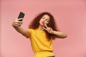 porträtt av attraktiv, rödhårig flicka med lockigt hår. bär gul t-shirt. framställning selfie på henne telefon. som visar fred tecken, vika ihop henne mun i en kyss. stå isolerat över pastell rosa bakgrund foto