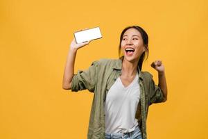 ung asiatisk dam visar tom smartphone -skärm med positivt uttryck, ler brett, klädd i vardagskläder som känner lycka på gul bakgrund. mobiltelefon med vit skärm i kvinnlig hand.
