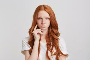 närbild av allvarlig sträng rödhårig ung kvinna med lång vågig hår bär t skjorta utseende arg och koncentrerad isolerat över vit bakgrund foto