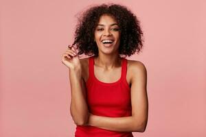 attraktiv glad afrikansk amerikan flicka med afro frisyr ser svälla underbar spelar med stå av lockigt mörk hår, skrattande, bär röd undertröja, isolerat på rosa bakgrund foto