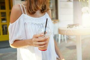 beskurna skott av skön ung kvinna bär vit sommar klänning innehav tös av milkshake gående nära utomhus- Kafé på de gata i de stad foto