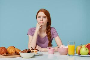 ledsen upprörd rödhårig flicka ser kamera med missnöje, tänker handla om diet, extra kalorier, bakning mat och färsk frukt juice yoghurt lägga på de tabell, på en blå bakgrund. friska mat begrepp foto