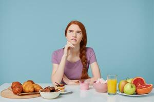 rödhårig flicka ser uppåt trodde handla om Vad till äta för frukost, sitter på en tabell på som lögner bakning Produkter och färsk frukt juice yoghurt, isolerat på en blå bakgrund. mat hälsa begrepp. foto