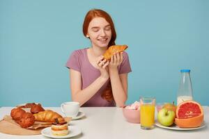 söt rödhårig flicka med en fläta sitter på en tabell, har frukost, med spänning nöje utseende på croissant, bakverk på de tabell och färsk frukt, på en blå bakgrund foto