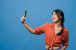 leende bedårande asiatisk kvinna som gör selfie -foto på smart telefon med positivt uttryck i vardagskläder och stativ isolerad på blå bakgrund. glad förtjusande glad kvinna jublar över framgång.