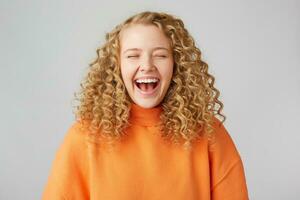 uthållig Lycklig glad lockigt hår blond stängd henne ögon med nöje, åtnjuter de ögonblick skrattande, bär en värma orange oversize Tröja, isolerat på vit bakgrund foto