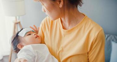 glad glad asiatisk familj mamma håller sjuk småbarn flicka i famnen och fäst den febernedsättande gelén på baby pannan promenad i vardagsrummet hemma. medicinsk vårdförsäkring, behandling och vårdkoncept. foto