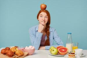 en flicka tänkande handla om rätt näring är Sammanträde på en tabell handla om till ha frukost, utseende eftertänksamt upp främre bakverk och friska mat, på henne huvud är innehav en röd äpple isolerat på blå bakgrund foto