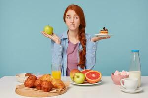 hård val. porträtt av skön kvinna påfrestande till välja mellan friska och ohälsosam mat medan isolerat på blå bakgrund. kvinna innehav kaka och äpple i händer foto