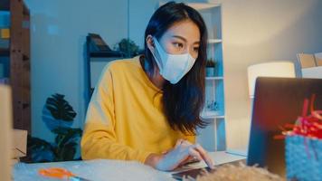 ung asiatisk affärskvinna bära ansiktsmask kontrollera inköpsorder och svara klient inkorg chatt på bärbar dator på hemmakontor på natten. småföretagare, onlinemarknadsleverans, livsstil frilansande koncept. foto