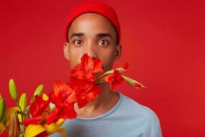 stänga upp av ung chockade kille i röd hatt och blå t-shirt, innehar en bukett i hans händer och täckt mun med blommor, utseende på de kamera med bred öppen ögon, står över röd bakgrund. foto