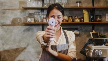 porträtt ung asiatisk kvinnlig restaurangpersonal som använder infraröd termometerchecker eller temperaturpistol på kundens panna innan de går in på urban caférestaurang. livsstil ny normal efter corona -virus. foto