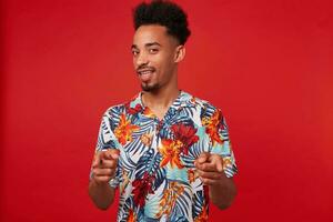 ung positiv afrikansk amerikan man bär i hawaiian skjorta, utseende på de kamera och brett ler, visar fingrar till de kamera och säger du Häftigt står över röd bakgrund. foto