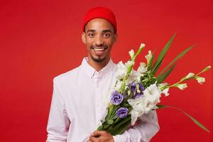 porträtt av ung Lycklig afrikansk amerikan man, bär i vit skjorta och röd hatt, utseende på de kamera och innehar bukett , står över röd bakgrund och leende. foto
