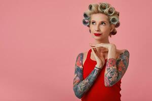 studio Foto av ung charmig blond kvinna med tatueringar ser positivt uppåt och hopfällbar Uppfostrad händer, har papiljotter på henne huvud medan Framställ över rosa bakgrund