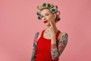 närbild av Söt ung blond tatuerade kvinna med papiljotter på henne huvud och röd mun ser listigt på kamera och leende lite, klädd i tillfällig kläder medan stående över rosa bakgrund foto