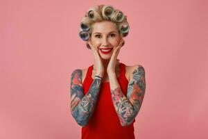 porträtt av skön ung Lycklig blond kvinna innehav palmer på henne kinder och leende allmänt medan Framställ över rosa bakgrund, har tatueringar på henne händer och papiljotter på henne huvud foto