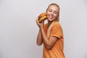 Lycklig ung lång håriga blond kvinna med tillfällig frisyr förvaring gott färsk hamburgare i henne händer och leende glatt med stängd ögon, stående mot vit bakgrund foto