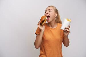 trevlig ser positiv ung blond lady med hästsvans frisyr förvaring ögon stängd medan njuter henne gott hamburgare, stående över vit bakgrund i tillfällig kläder foto