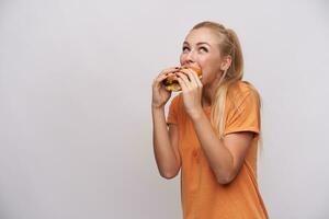 inomhus- Foto av ung glad blond kvinna med tillfällig frisyr äter hamburgare med bra nöje medan Framställ över vit bakgrund i orange t-shirt