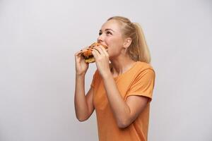 glad hungrig ung blond kvinna med tillfällig frisyr bitande ivrigt av stor färsk burger medan stående över vit bakgrund, klädd i orange t-shirt foto