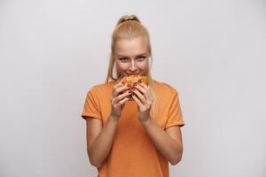 nöjd ung attraktiv blond kvinna med hästsvans förtjusande färsk burger och ser glatt på kamera, bär orange t-shirt medan stående mot vit bakgrund foto