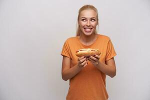 upphetsad Söt ung lång håriga blond lady med varm hund i Uppfostrad händer ser åt sidan med upprörd leende och försmak utsökt middag, stående över vit bakgrund i orange t-shirt foto