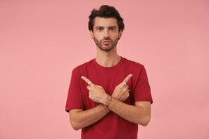 studio Foto av mörk håriga manlig med trendig frisyr Framställ över rosa bakgrund i röd t-shirt, ser till kamera med lugna ansikte och vikta mun, som visar med pekfingrar i annorlunda sidor