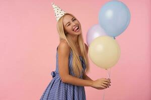 porträtt av glad ung lång håriga blond kvinna i blå sommar klänning och kon hatt Framställ över rosa bakgrund med flerfärgad luft ballonger i händer, blinka på kamera och som visar tunga lyckligt foto