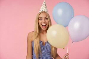 glad Söt lång håriga blond lady har glad stunder i henne liv under födelsedag fest, Framställ över rosa bakgrund med flerfärgad luft ballonger, varelse i hög anda foto