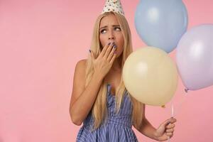 uttråkad Söt ung kvinna med lång blond hår bär festlig kläder och kon hatt, innehav knippa av helium ballonger medan Framställ över rosa bakgrund, gapande och beläggning mun med Uppfostrad hand foto