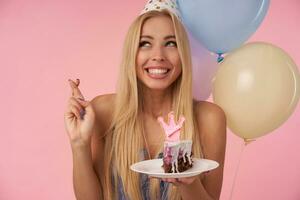 glad ung lång håriga kvinna med lång blond hår korsning fingrar medan framställning önskar på henne födelsedag, innehav bit av kaka med ljus över flerfärgad luft ballonger och rosa bakgrund foto