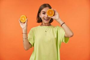 Tonårs flicka, glad och Lycklig med kort brunett hår innehav apelsiner över henne öga, omslag ett öga. stående över orange bakgrund. bär grön t-shirt, tänder tandställning och armband foto