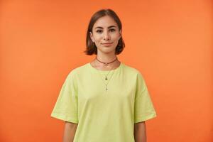 kvinna studerande, attraktiv ung lady med kort brunett hår tittar på med förtroende på de kamera isolerat över orange bakgrund. bär grön t-shirt, halsband och örhängen. foto